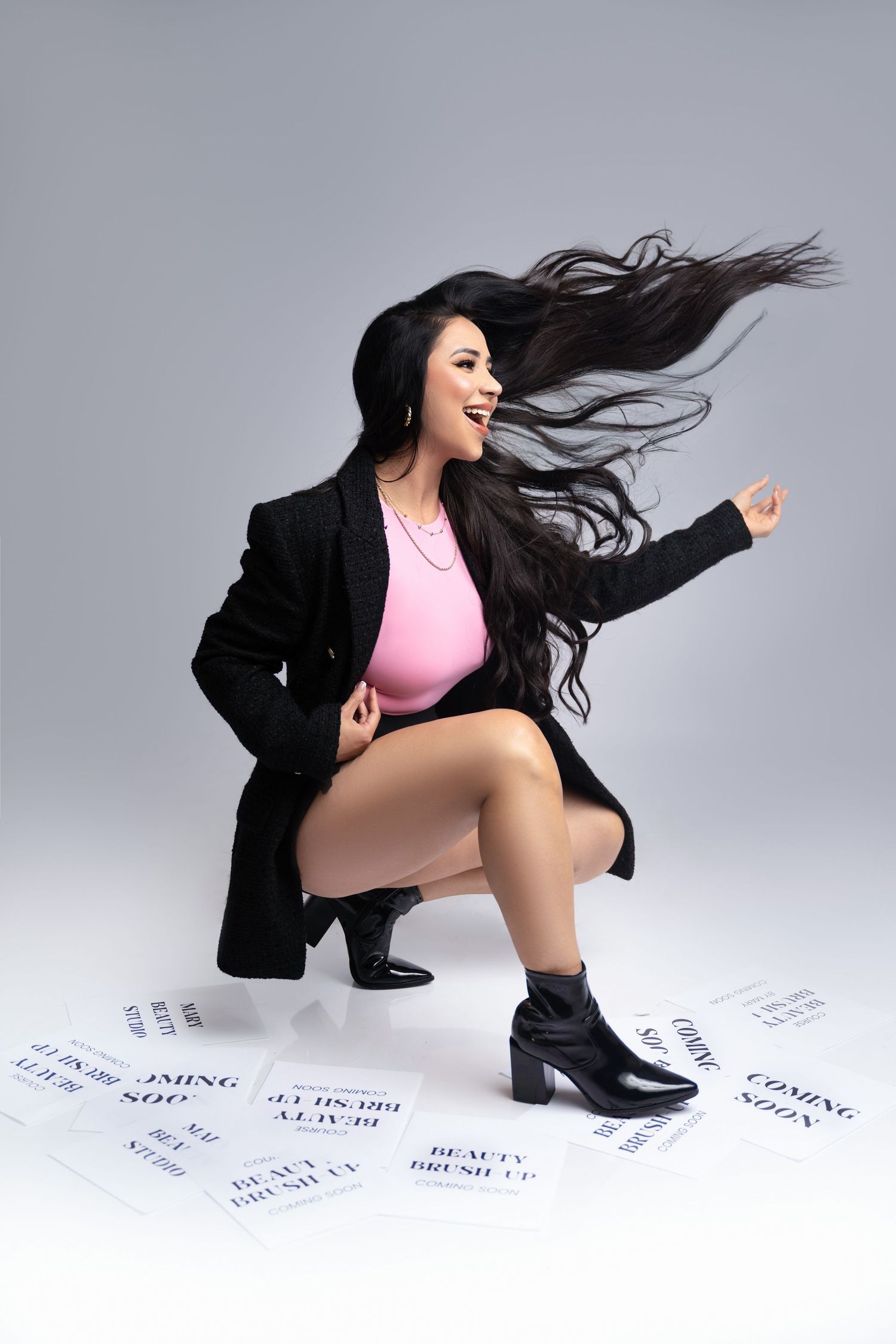 Woman in black blazer and pink top crouching with flowing hair, surrounded by beauty event flyers on gray background.
