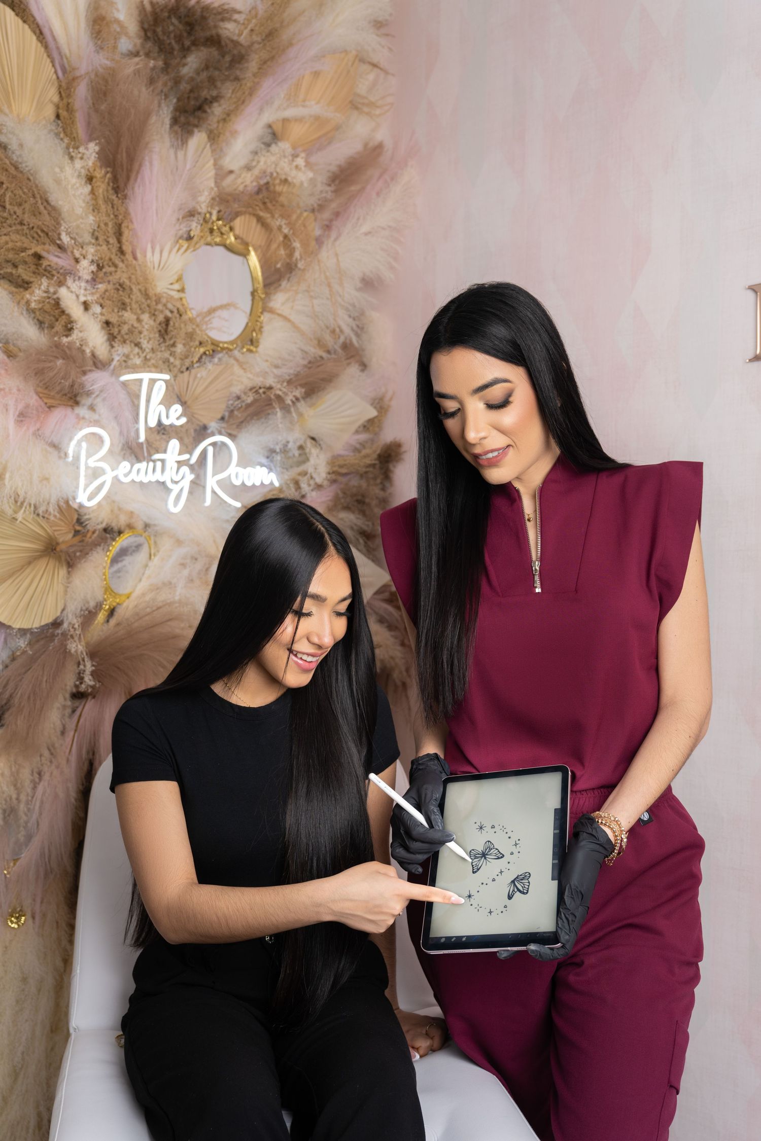 Two women at The Beauty Room salon review a tablet displaying beauty designs in an elegant pink studio setting.