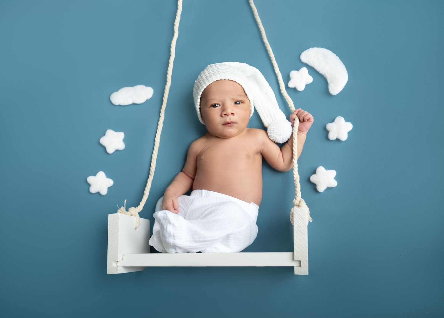 Newborn baby in white diaper and knit hat resting on a white swing with stars and moon on blue background.