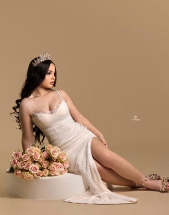 Young woman in white dress and tiara poses elegantly with pink roses on white pedestal against beige background.