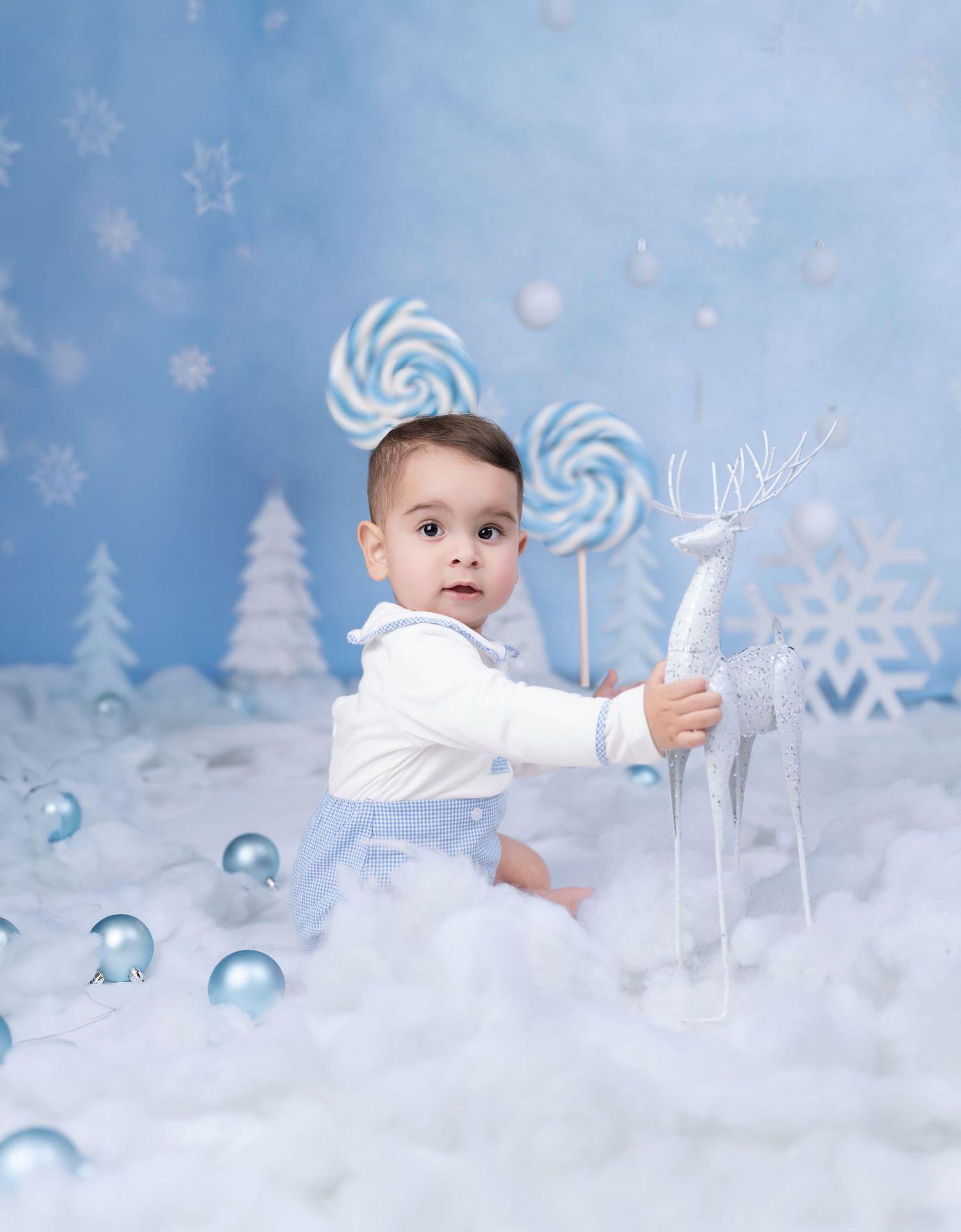 Baby boy in white outfit holds reindeer figurine in winter wonderland scene with blue ornaments and lollipops.
