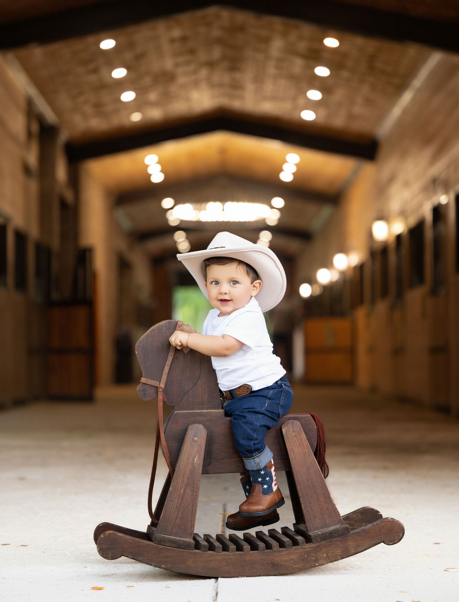 Adorable toddler in cowboy hat and boots riding a wooden rocking horse inside a warmly lit barn aisle.