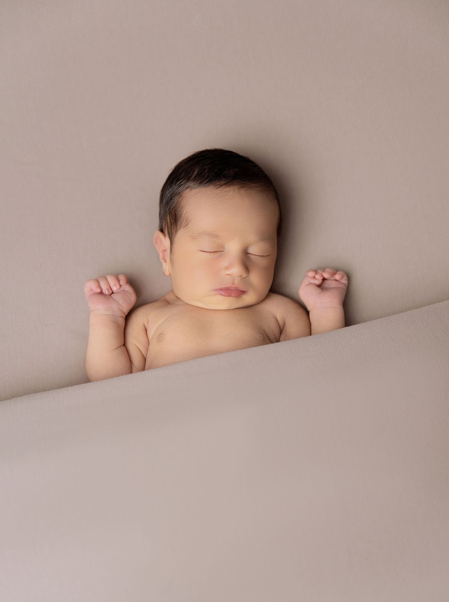 Peaceful sleeping newborn baby with dark hair, tiny fists raised, lying on a soft beige blanket in a studio portrait.