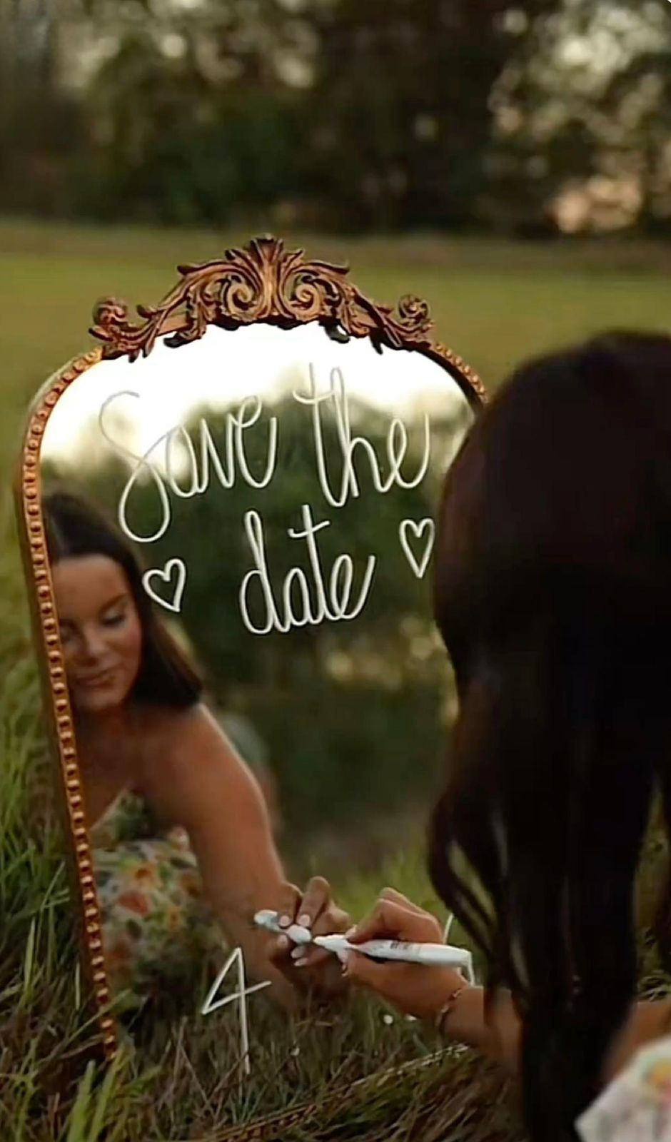 Woman writes save the date on ornate gold mirror outdoors in natural setting.