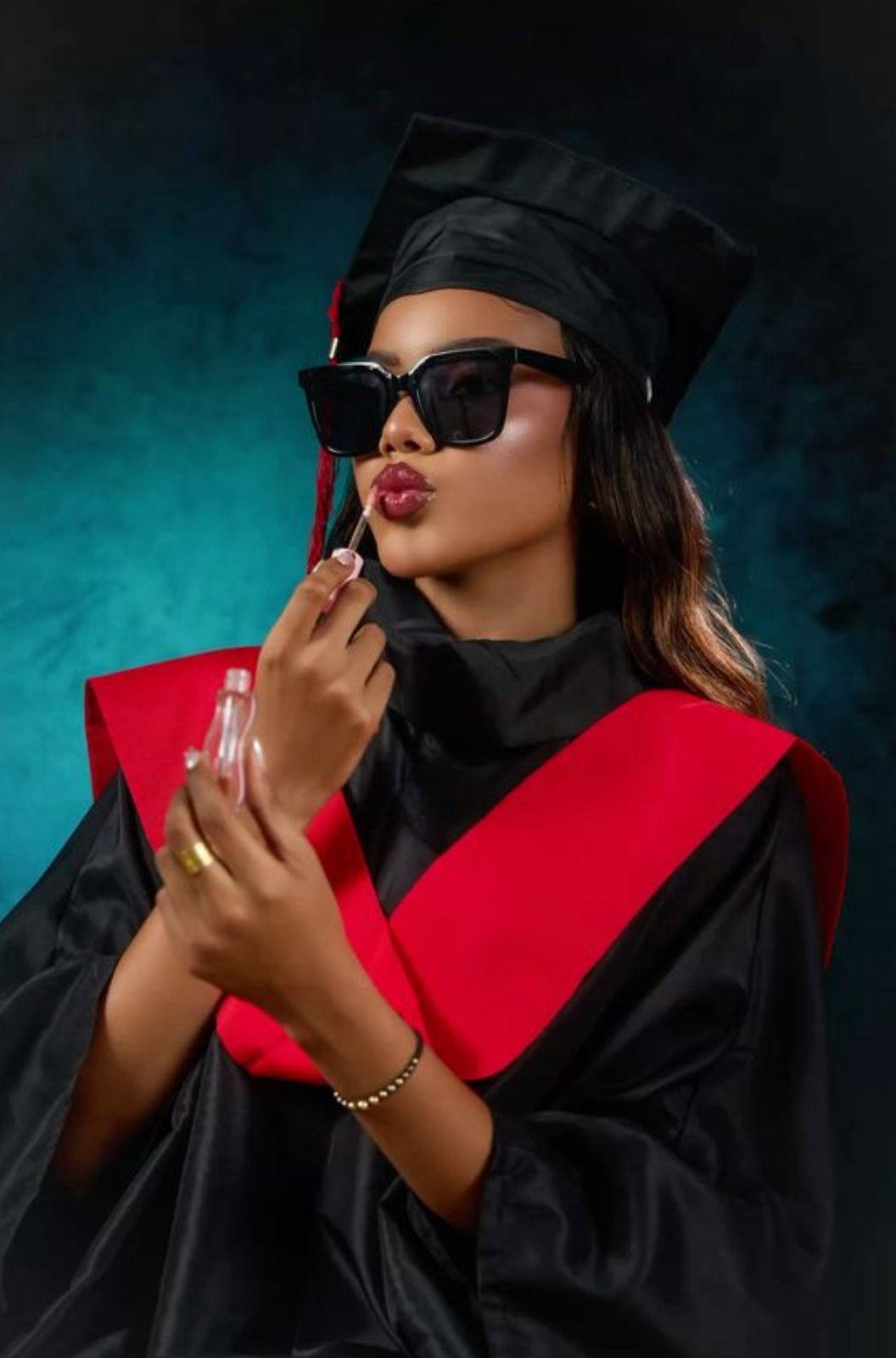 Graduate in black and red academic regalia wearing sunglasses applies lipstick against turquoise backdrop.