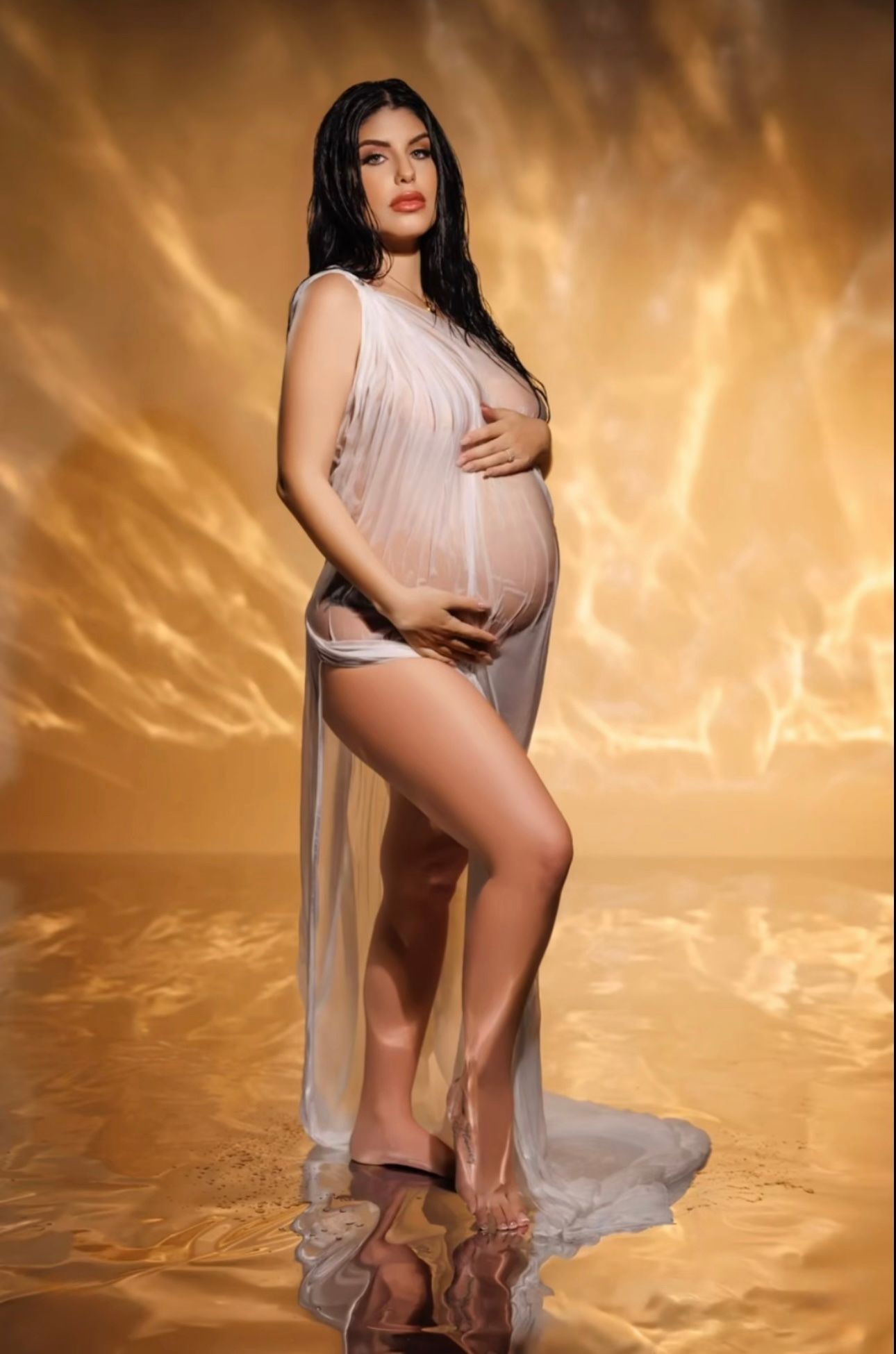 Pregnant woman in sheer white gown poses gracefully against a golden glowing backdrop in a maternity photoshoot.