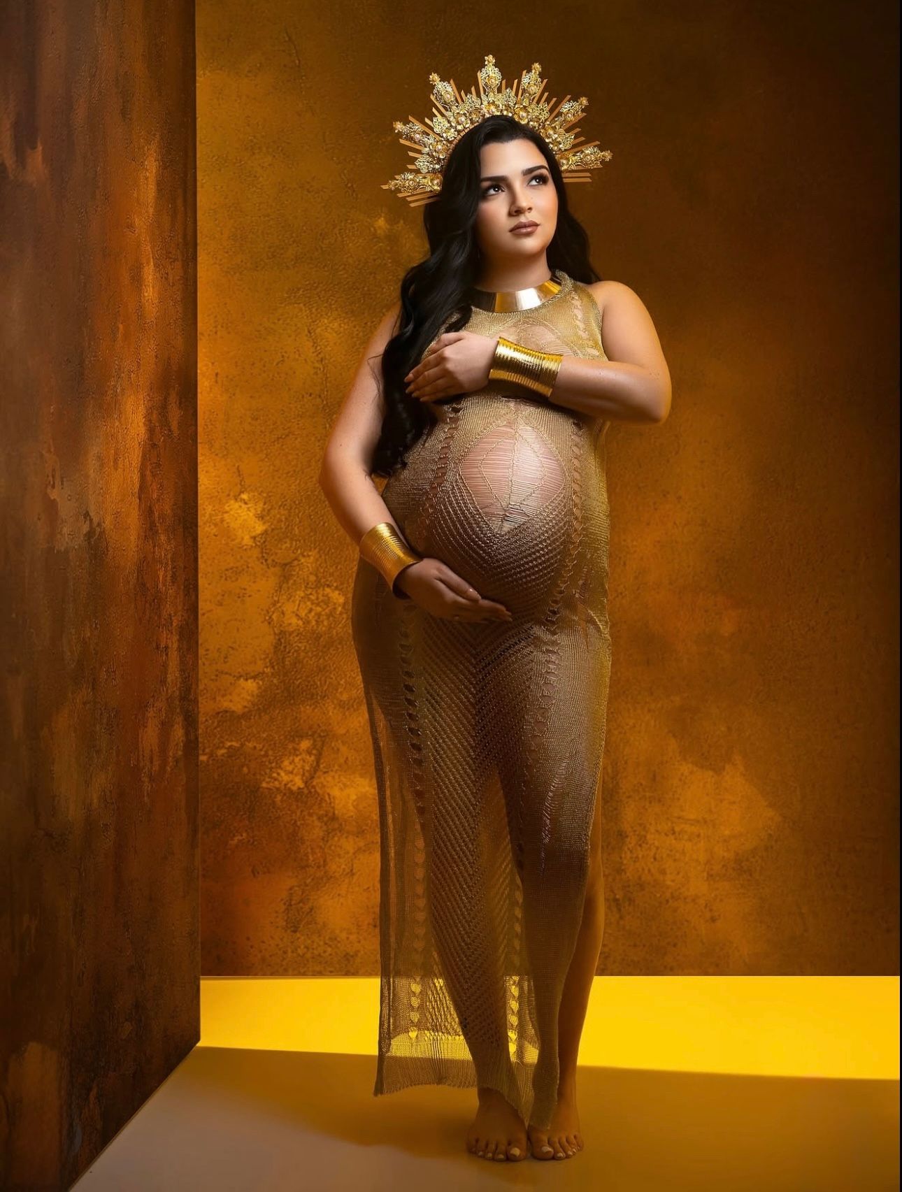 Pregnant woman in gold mesh gown and crown poses regally against golden backdrop in elegant maternity photo shoot.