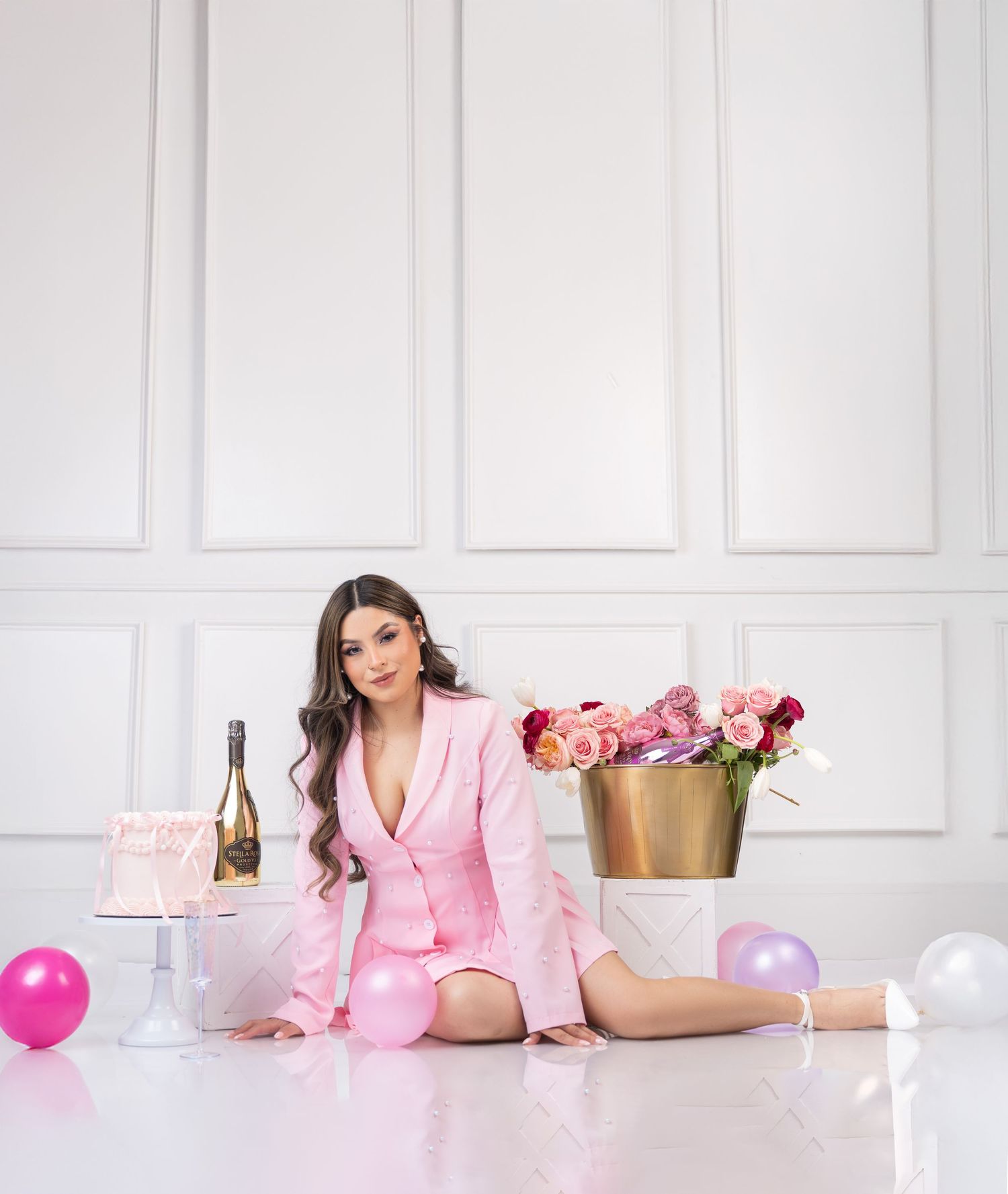 A person in pink loungewear poses on white floor with champagne, balloons and roses in an elegant room setting.