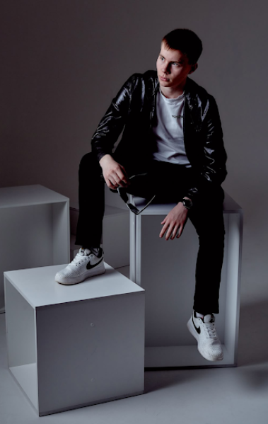 Stylish model in black leather jacket and white sneakers poses on geometric white blocks against dark backdrop.