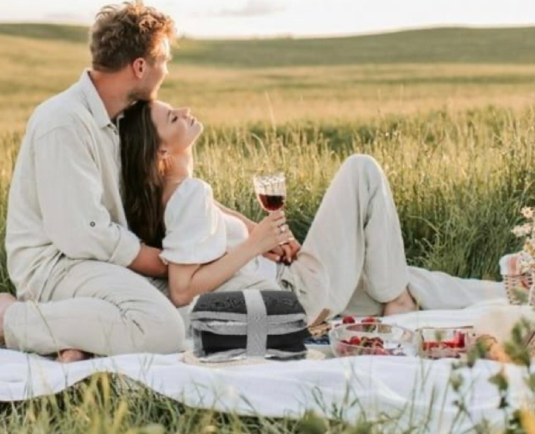 A couple enjoys a romantic sunset picnic in a golden wheat field with wine and snacks laid out on a white blanket.