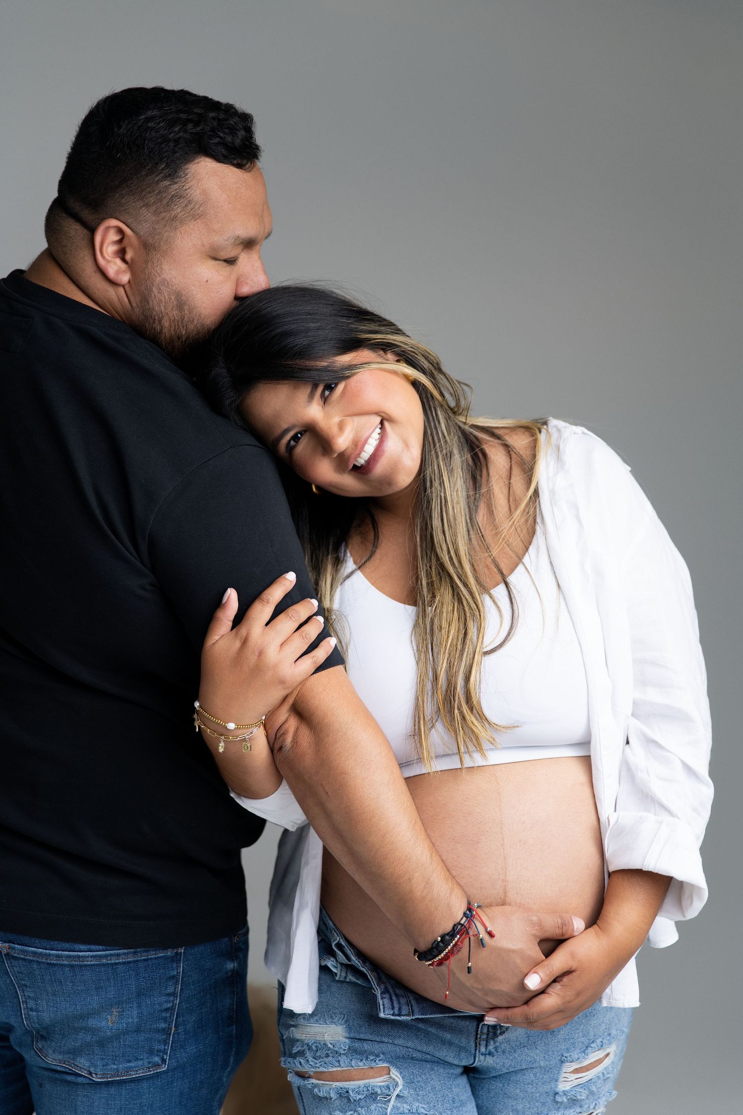 A couple shares tender moments during a maternity photoshoot against a grey studio backdrop.