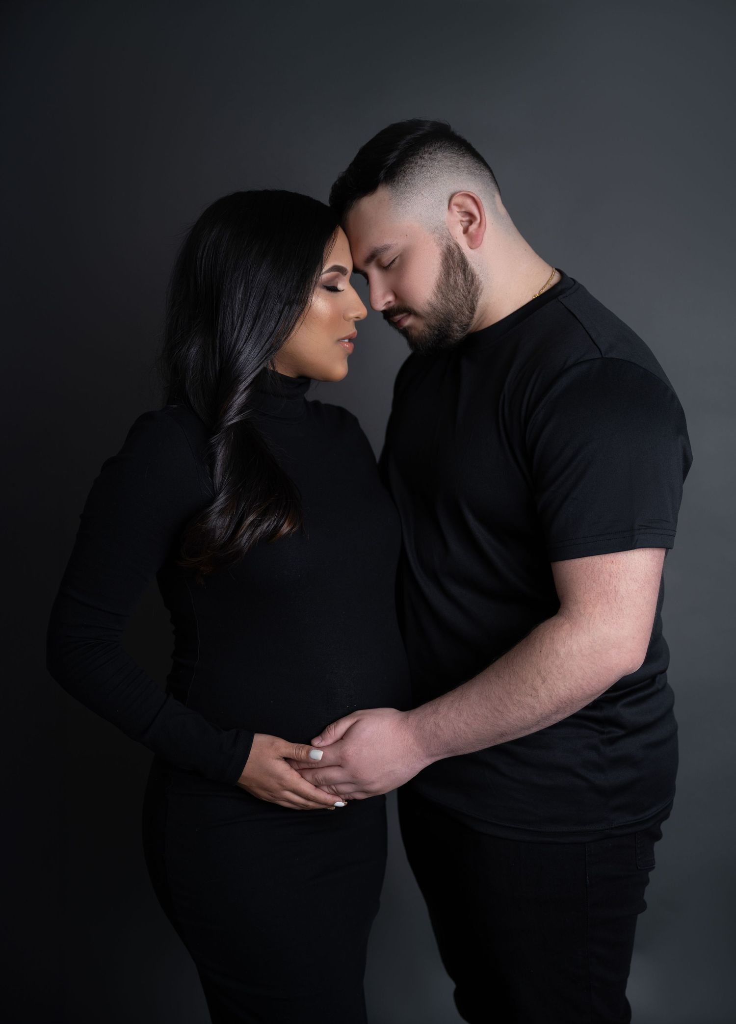 Intimate maternity portrait of a couple in black outfits against dark backdrop.