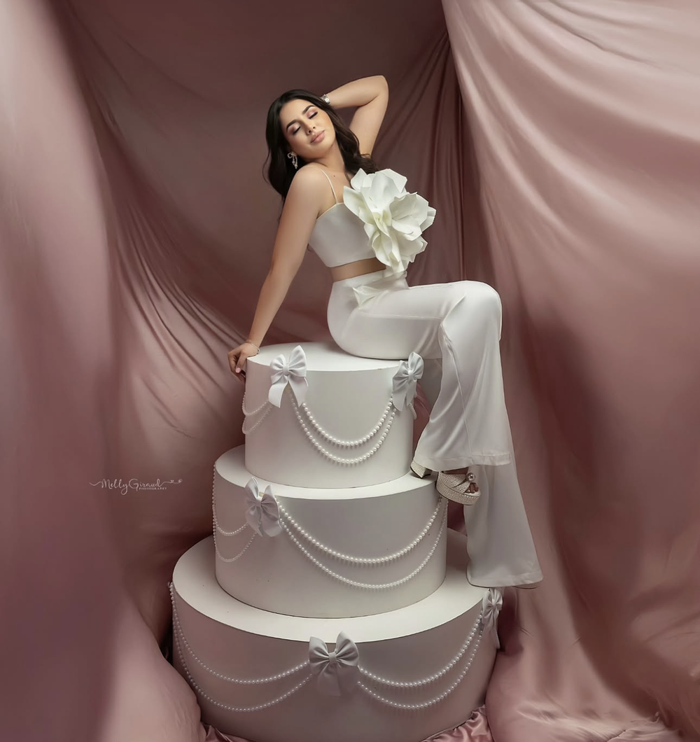 Woman in white outfit posing elegantly beside a large tiered white wedding cake against a pink backdrop.