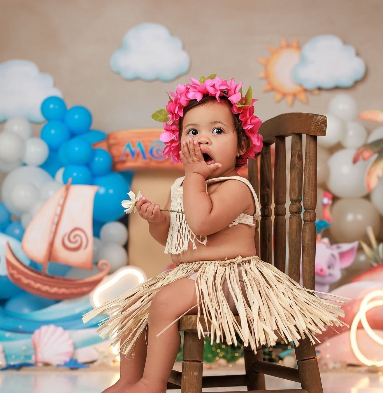 Baby girl in hula costume with floral crown sits on chair at Moana-themed first birthday cake smash photo shoot.