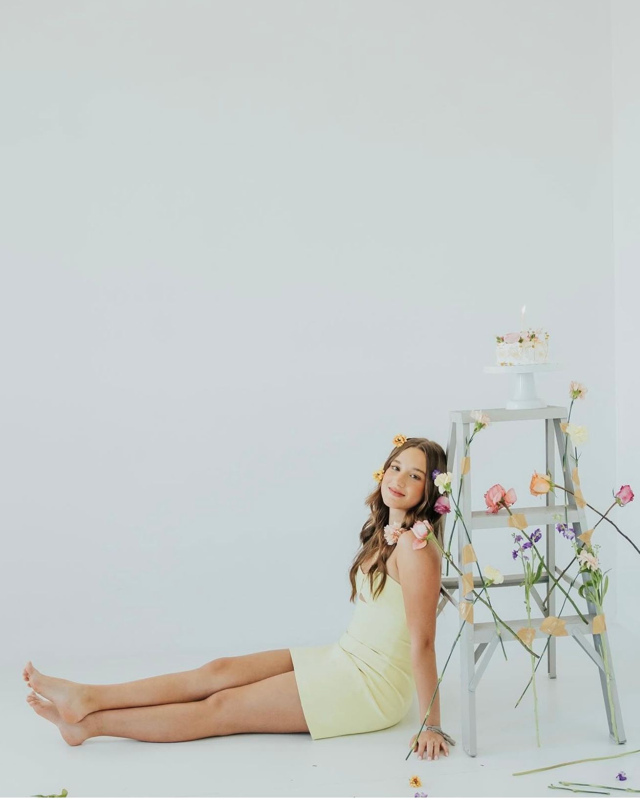 Young girl in yellow romper sitting on white floor beside ladder decorated with colorful spring flowers.