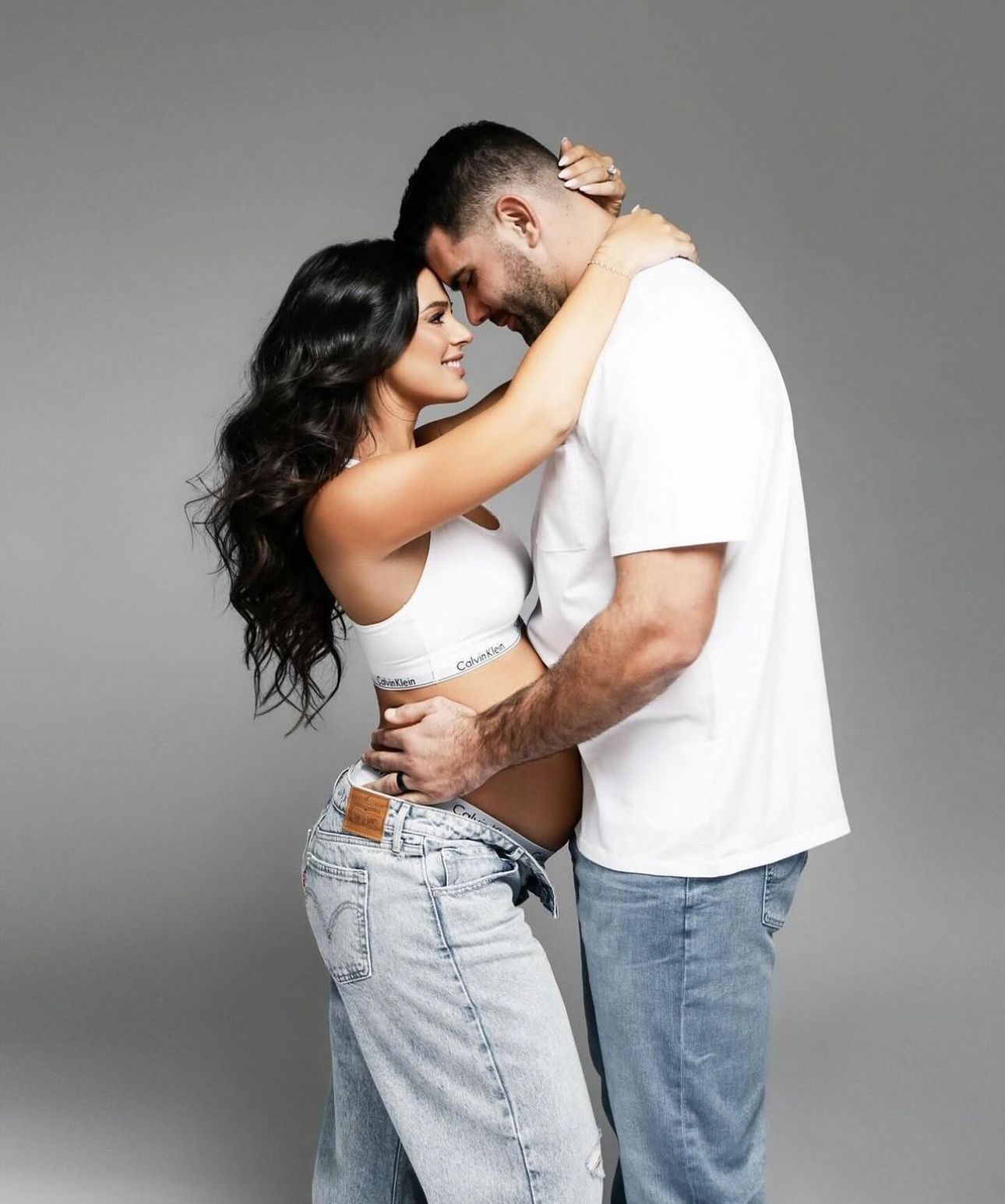 Couple embracing intimately in white tops and jeans against gray background, woman with long dark hair.