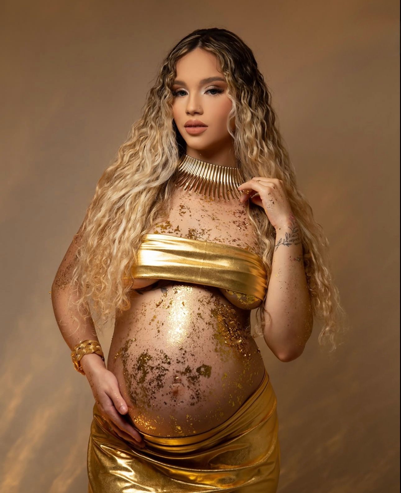 Pregnant woman in gold metallic outfit with body glitter, long wavy blonde hair, posing for maternity photoshoot.