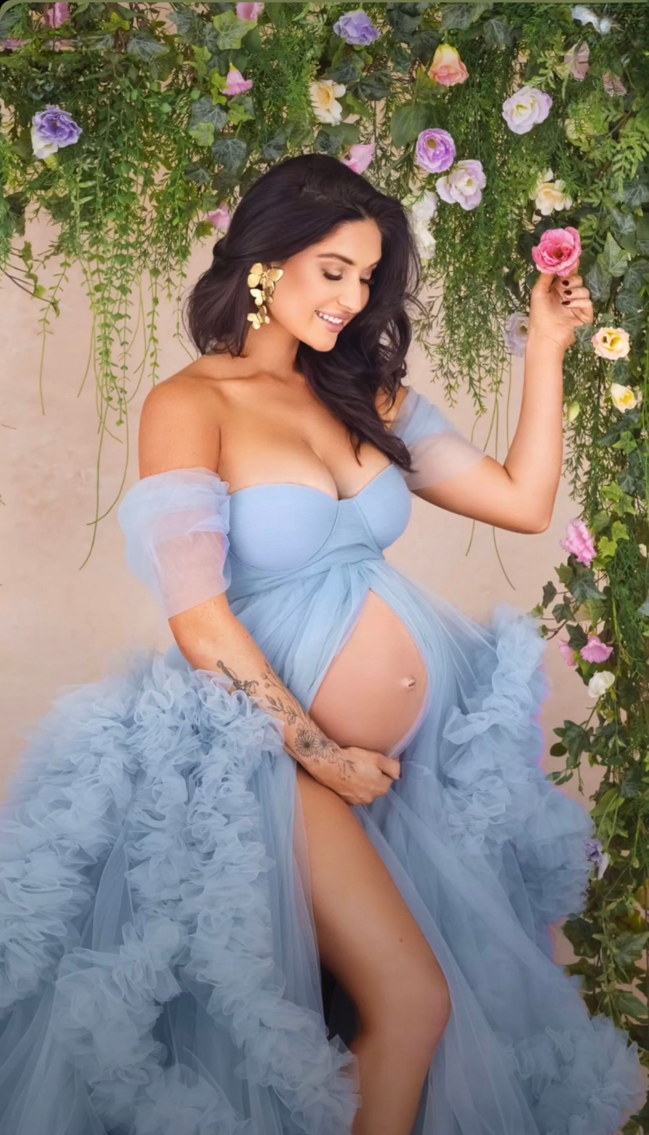 Pregnant woman in a blue off-shoulder tulle gown posing with hanging floral backdrop.