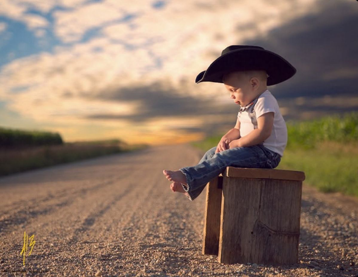 A toddler in a black cowboy hat sits on a wooden box along a rural road at sunset.