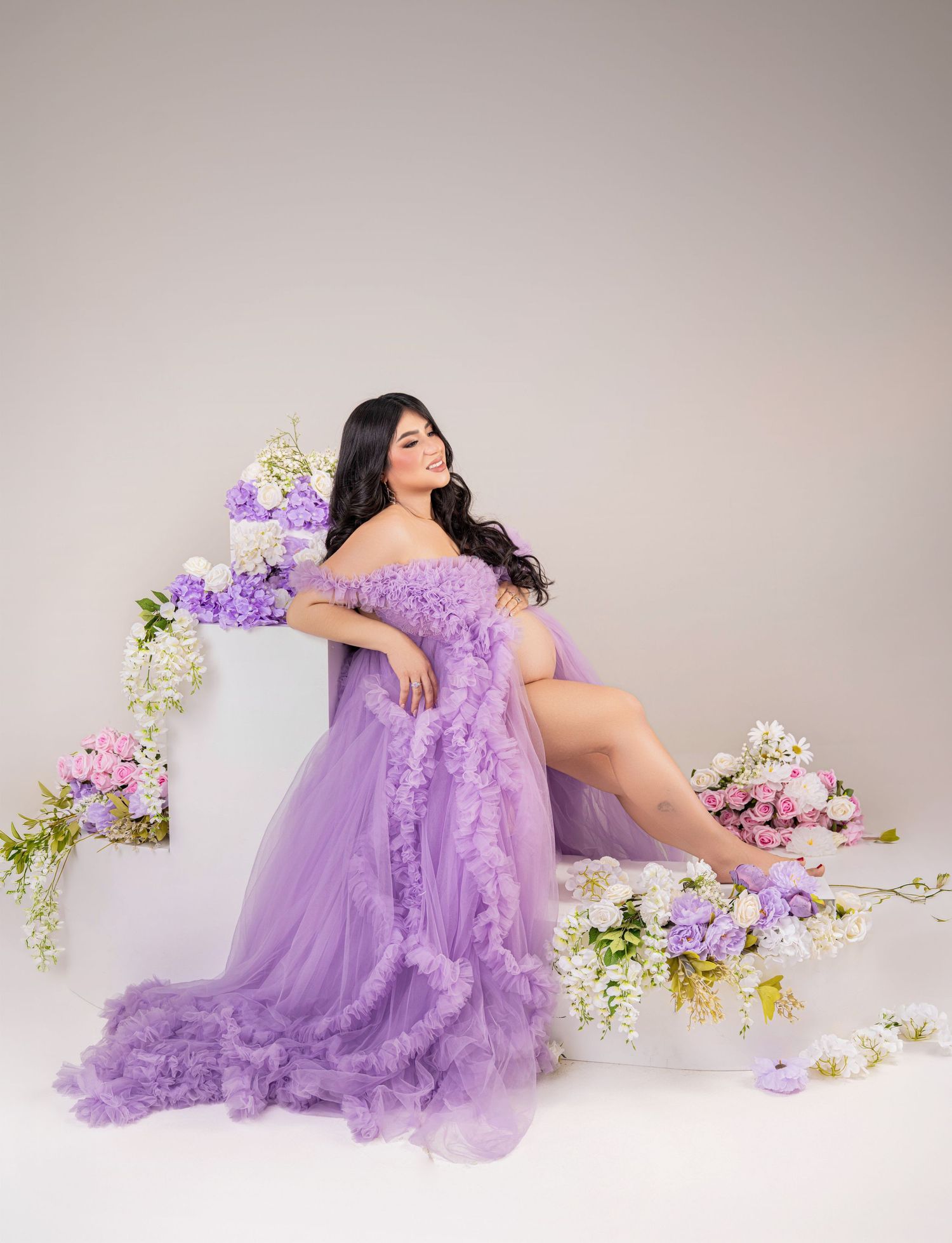 Woman in lavender ruffle tulle gown poses elegantly among pink and purple floral arrangements in studio.
