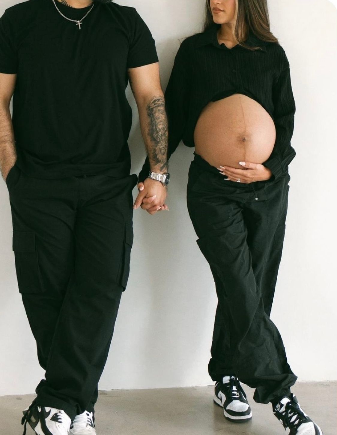 Pregnant couple in matching black outfits holding hands, woman cradling baby bump, both wearing black and white Nike sneakers.