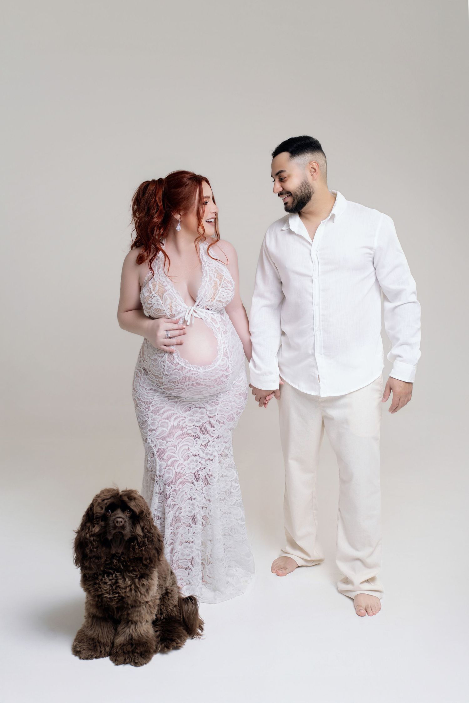 A couple in white attire poses together with a fluffy brown dog in a minimalist studio setting.