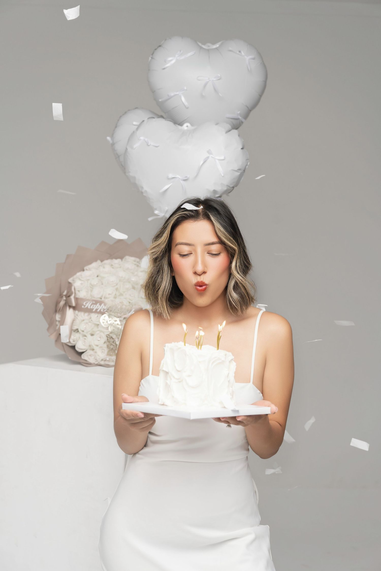 A person in a white dress blows out birthday candles on a cake while white heart balloons float above.