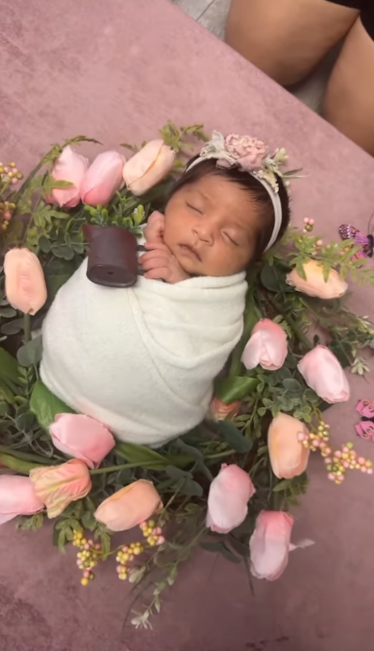 Newborn baby wrapped in white blanket nestled among pink tulips and greenery on pink backdrop.