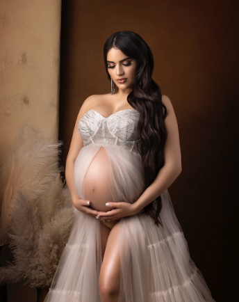 A pregnant woman in a white lace and tulle gown poses elegantly against a brown backdrop in a maternity photoshoot.