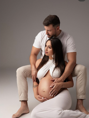 An intimate maternity photoshoot with couple in white and neutral tones against a grey studio backdrop.