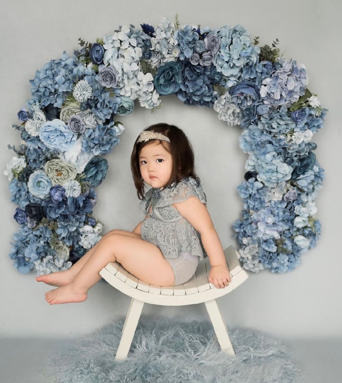 Baby in a white dress poses with delicate blue and white floral wreaths on a rocking horse bench.