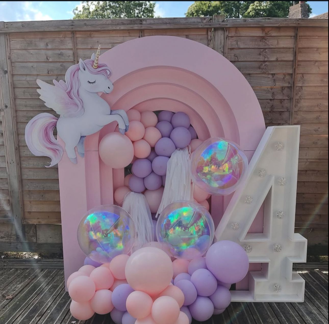 A pink and purple balloon arch with a unicorn decoration and number 4 display against a wooden fence backdrop.