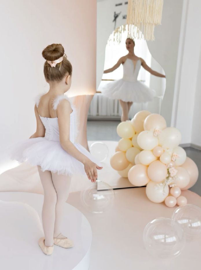 A ballet dancer in white tutu practices gracefully in a bright studio with soft peach-colored balloons in the background.