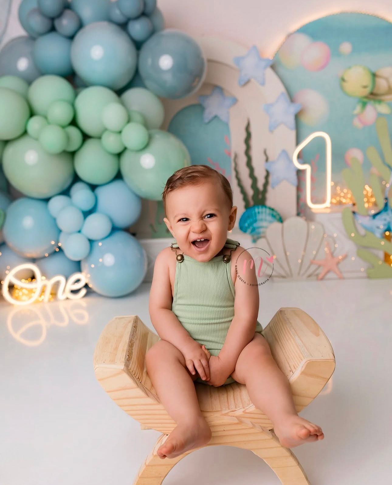 Baby in mint green romper laughs joyfully during first birthday photoshoot with pastel balloon garland backdrop.