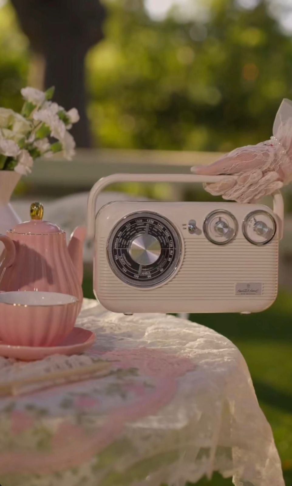 Vintage cream radio on floral lace tablecloth beside pink tea set and flowers in a garden setting.