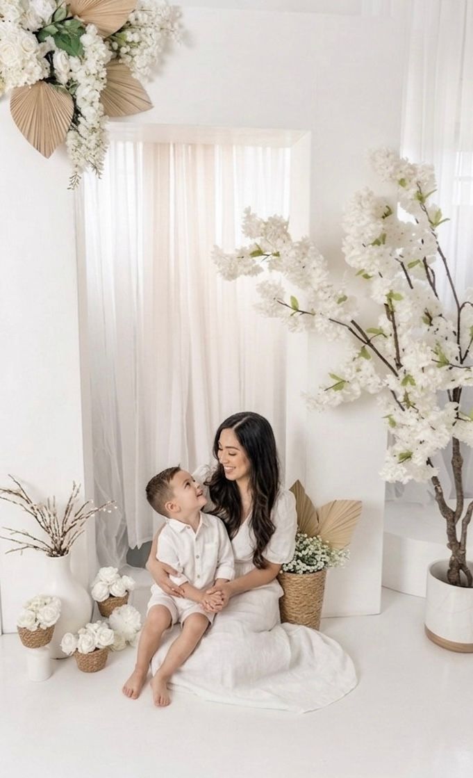 A mother and young child sit together in a white floral studio setting, both dressed in white, sharing a tender moment.