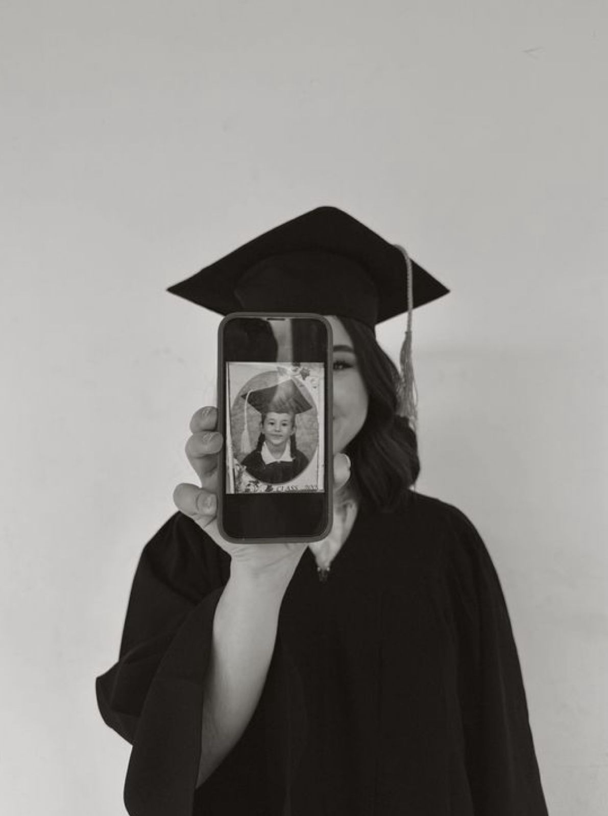 A graduate in black cap and gown holds up a phone displaying a photo, creating a recursive self-portrait effect.