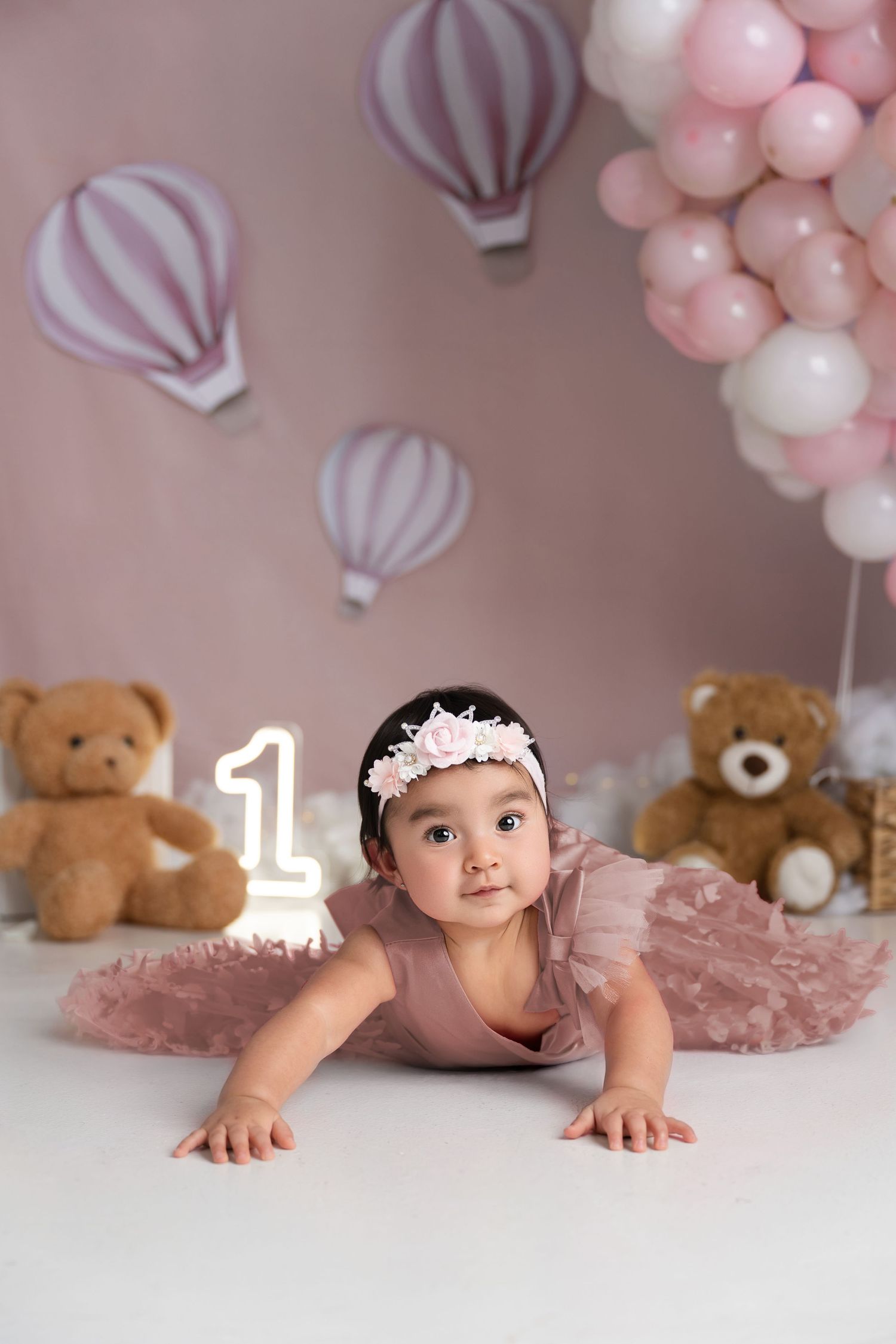 Baby girl in pink tutu celebrates first birthday with teddy bears, balloons, and number one candle in a dreamy pink setting.