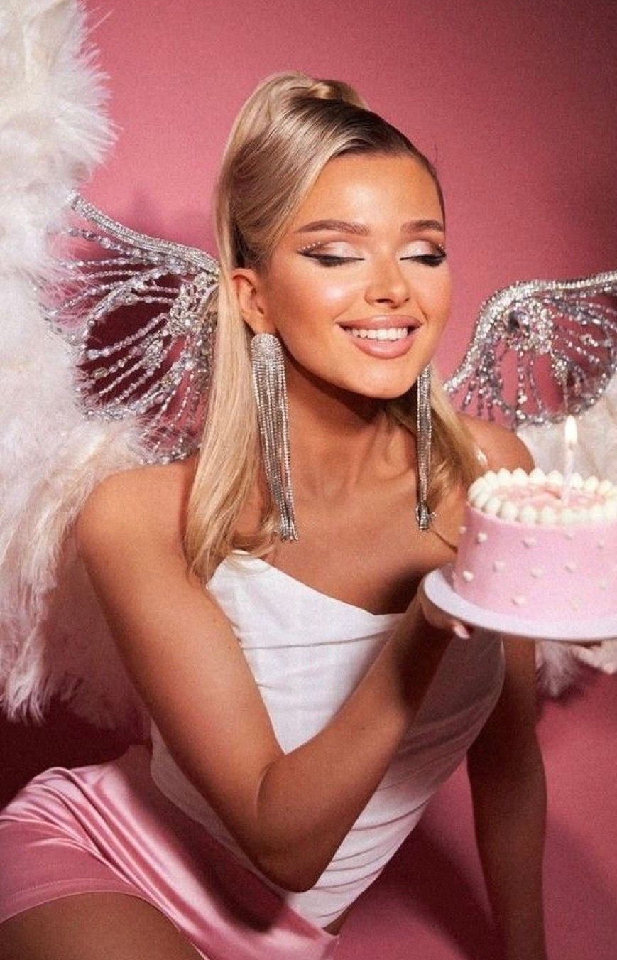 Blonde woman in white dress with sparkly angel wings holding a pink birthday cake on a pink background.