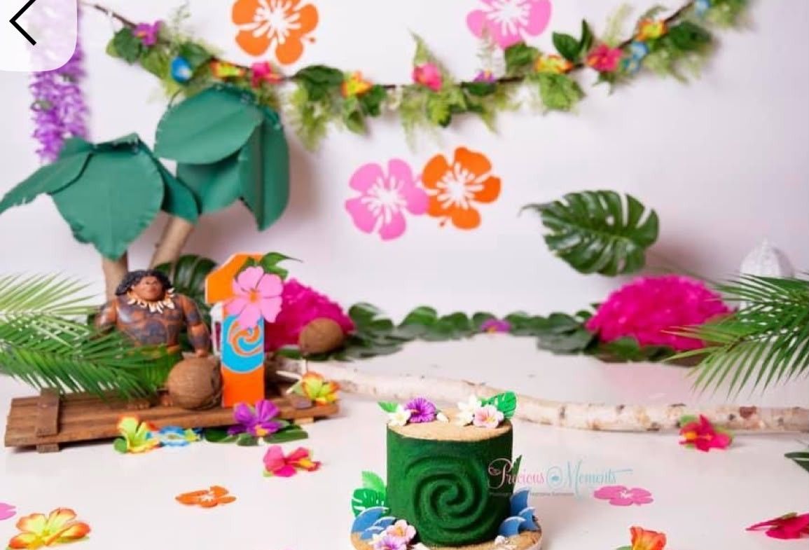 Moana-themed cake smash setup with tropical flowers, palm leaves, green spiral cake, and character figurines on white backdrop.