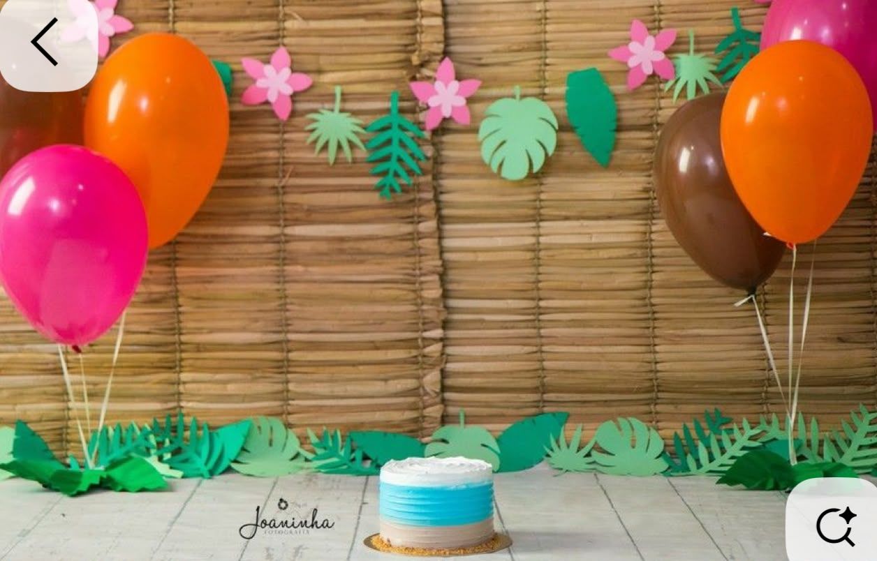 Tropical birthday party setup with colorful balloons, paper palm leaves, floral garland, and a small cake against a bamboo backdrop.