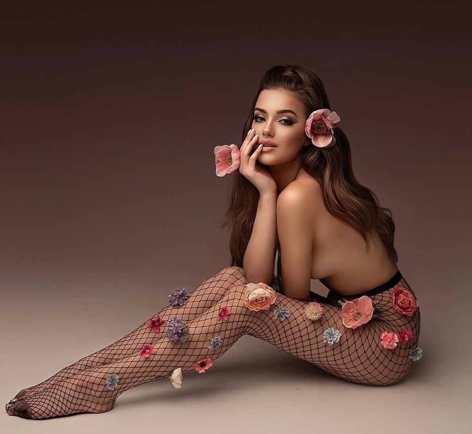 Woman in floral fishnet stockings poses gracefully with flowers in her hair against a warm studio background.