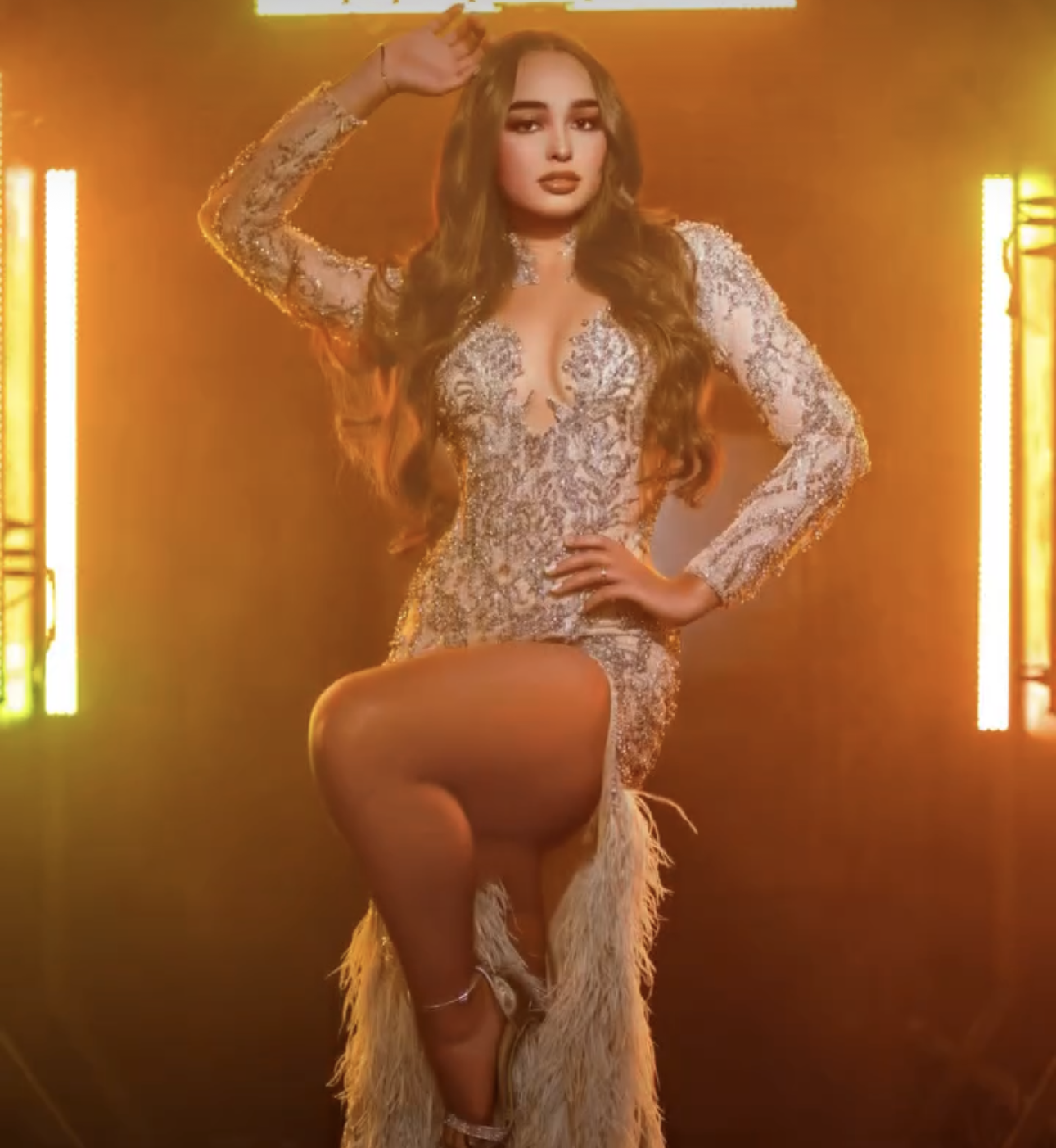 Woman in a beaded white long-sleeve mini dress with feather hem poses confidently under warm golden lights.