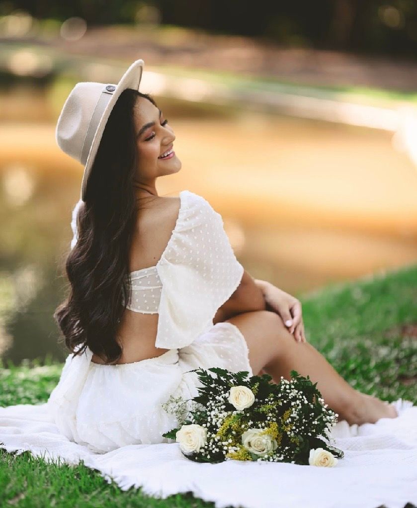 Smiling woman in white crop top and skirt with hat sits on blanket outdoors holding white roses bouquet.
