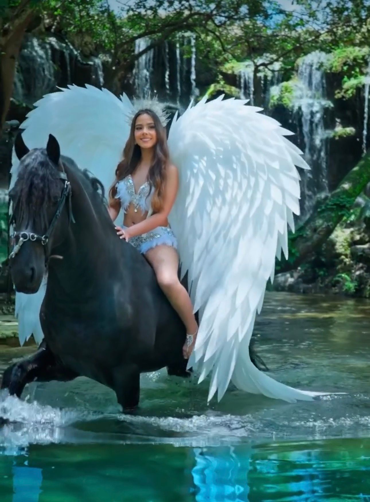 Young woman with angel wings rides a black horse through a serene waterfall pool in a fantasy-themed photo.