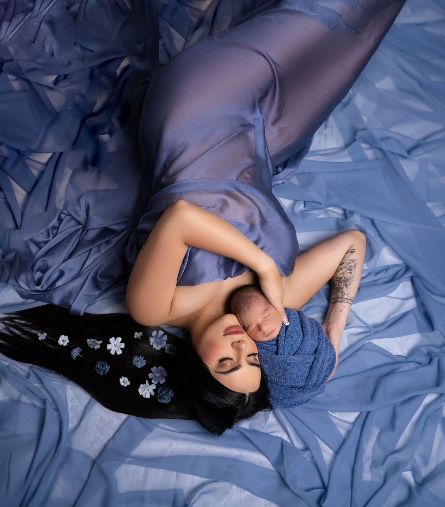 A tattooed woman in a blue gown lies on flowing fabric, cradling a newborn baby wrapped in blue cloth.