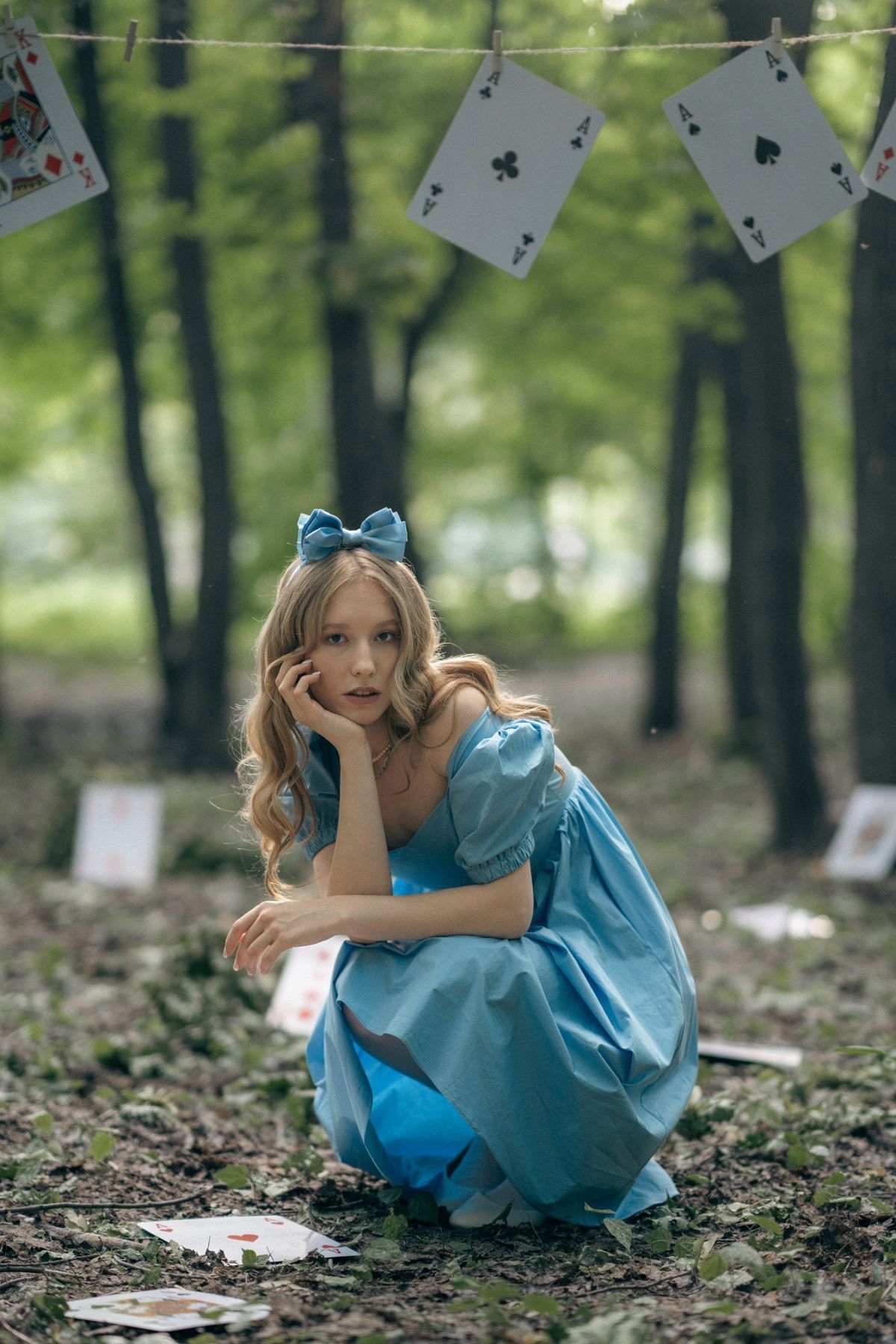 Young woman in blue Alice in Wonderland costume crouching in forest with playing cards hanging above.