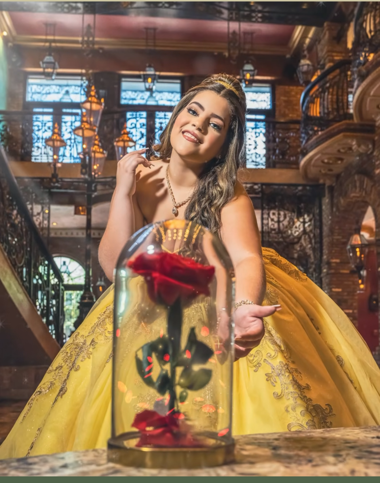 Young woman in yellow ball gown poses with enchanted rose under glass dome in ornate castle setting.