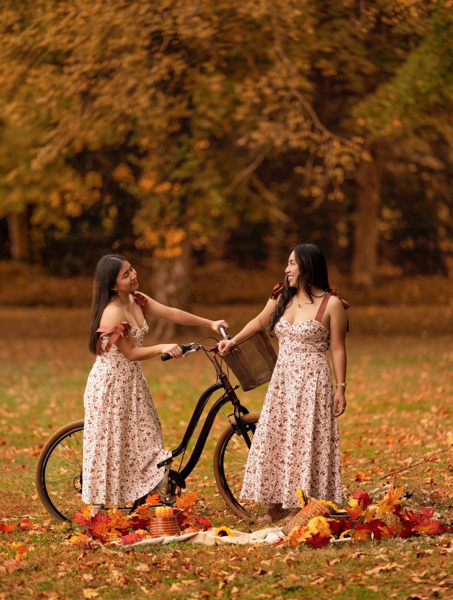 Two women in floral dresses pose with a vintage bicycle among autumn leaves in a golden park.