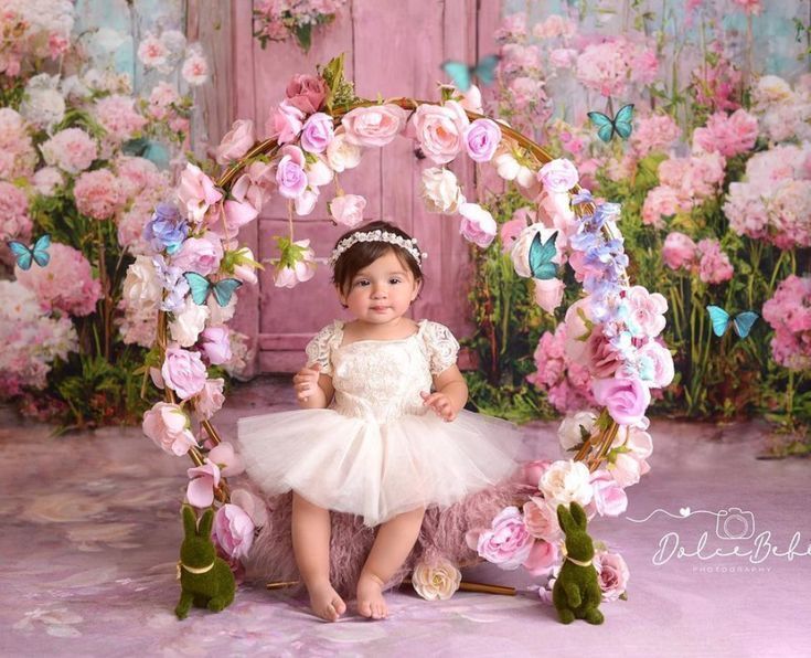 Baby girl in white dress sits on floral arch swing with pink roses and butterflies in a garden-themed photo shoot.